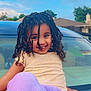 child, smiling, curly_hair, braids, yellow_shirt, purple_pants, car, hood, outdoor, daylight, suburban, house, sky, happy, playful, portrait, clothing, person, nature, casual