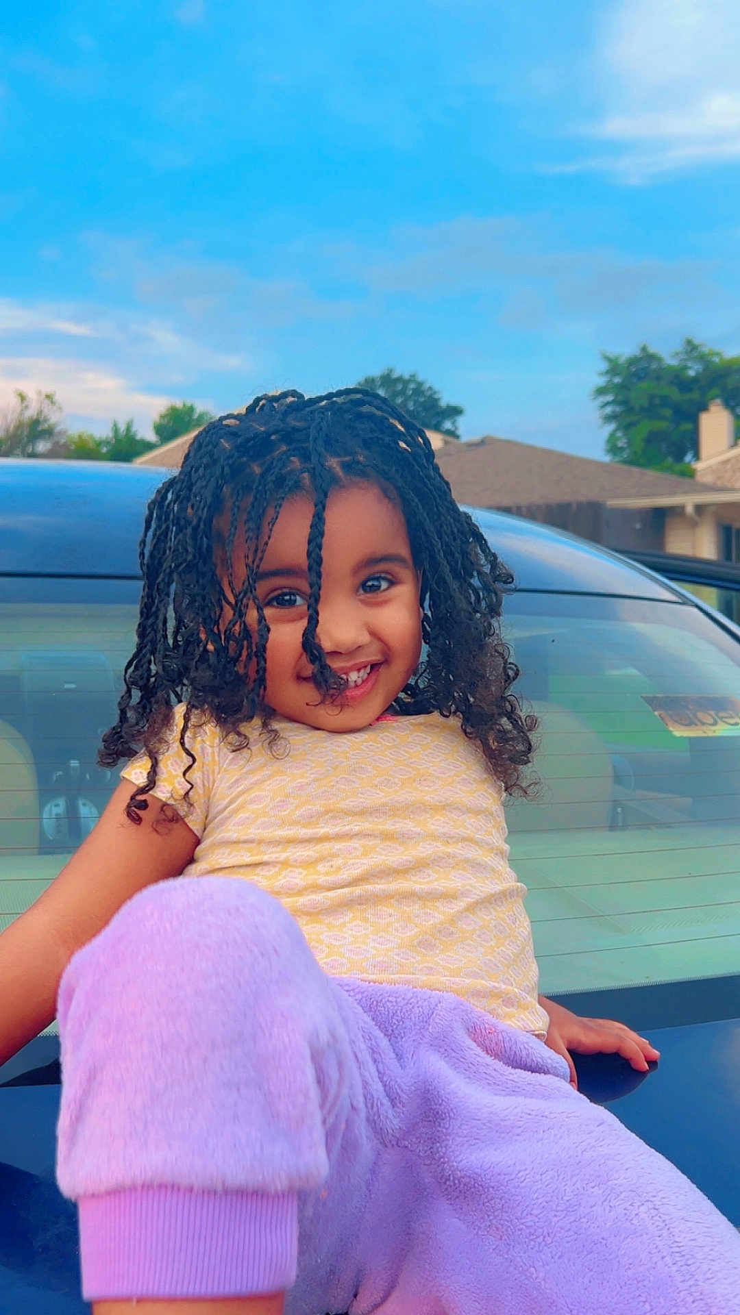 Hierani joined the competition — help win amazing prizes! child, smiling, curly_hair, braids, yellow_shirt, purple_pants, car, hood, outdoor, daylight, suburban, house, sky, happy, playful, portrait, clothing, person, nature, casual