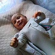 Eli joined the competition — help win amazing prizes! baby, infant, smile, face, onesie, blanket, cushion, bed, happy, child, portrait, indoor, young, cute, fist, clothing, person, lying_down, head, skin