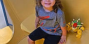 Osmar joined the competition — help win amazing prizes! child, curly_hair, grey_shirt, black_shorts, sneakers, toy, yellow_bench, indoor, smiling, happy, seated, playful, casual_clothing, young_child, person, portrait, colorful_toy, curly_hair_child, relaxed, daylight