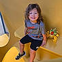 child, curly_hair, grey_shirt, black_shorts, sneakers, toy, yellow_bench, indoor, smiling, happy, seated, playful, casual_clothing, young_child, person, portrait, colorful_toy, curly_hair_child, relaxed, daylight