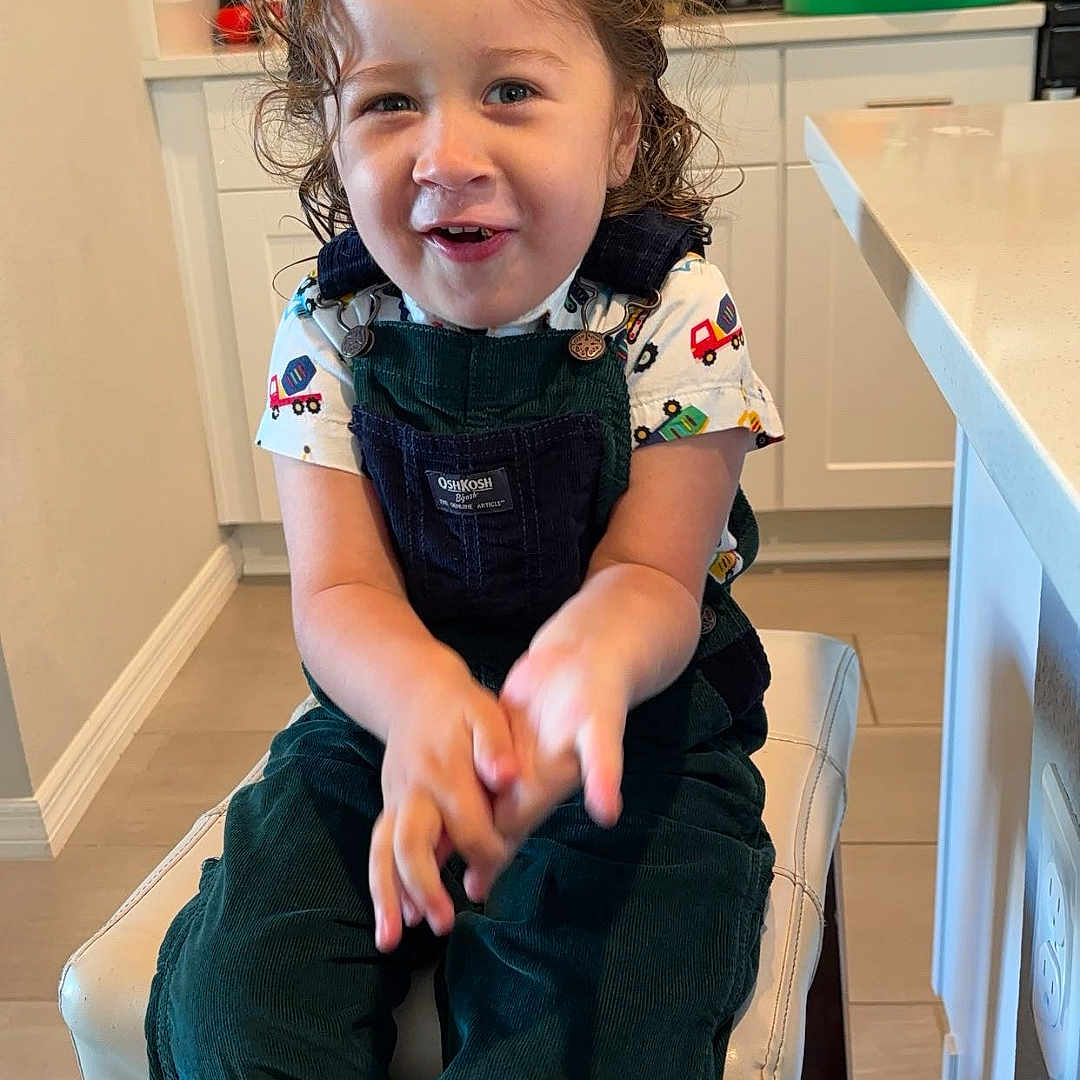 Osmar is registered to the contest to win money with this photo: appliance, cabinet, child, clapping, countertop, cupboard, curly_hair, floor, furniture, green_overalls, happy, indoor, kitchen, light, person, shirt, smiling, stool, toddler, toy_truck_pattern
