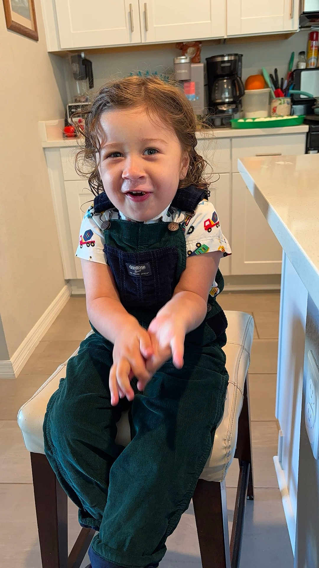 Osmar is registered to the contest to win money with this photo: child, toddler, smiling, curly_hair, green_overalls, kitchen, stool, clapping, happy, indoor, person, cabinet, countertop, appliance, cupboard, floor, shirt, toy_truck_pattern, furniture, light
