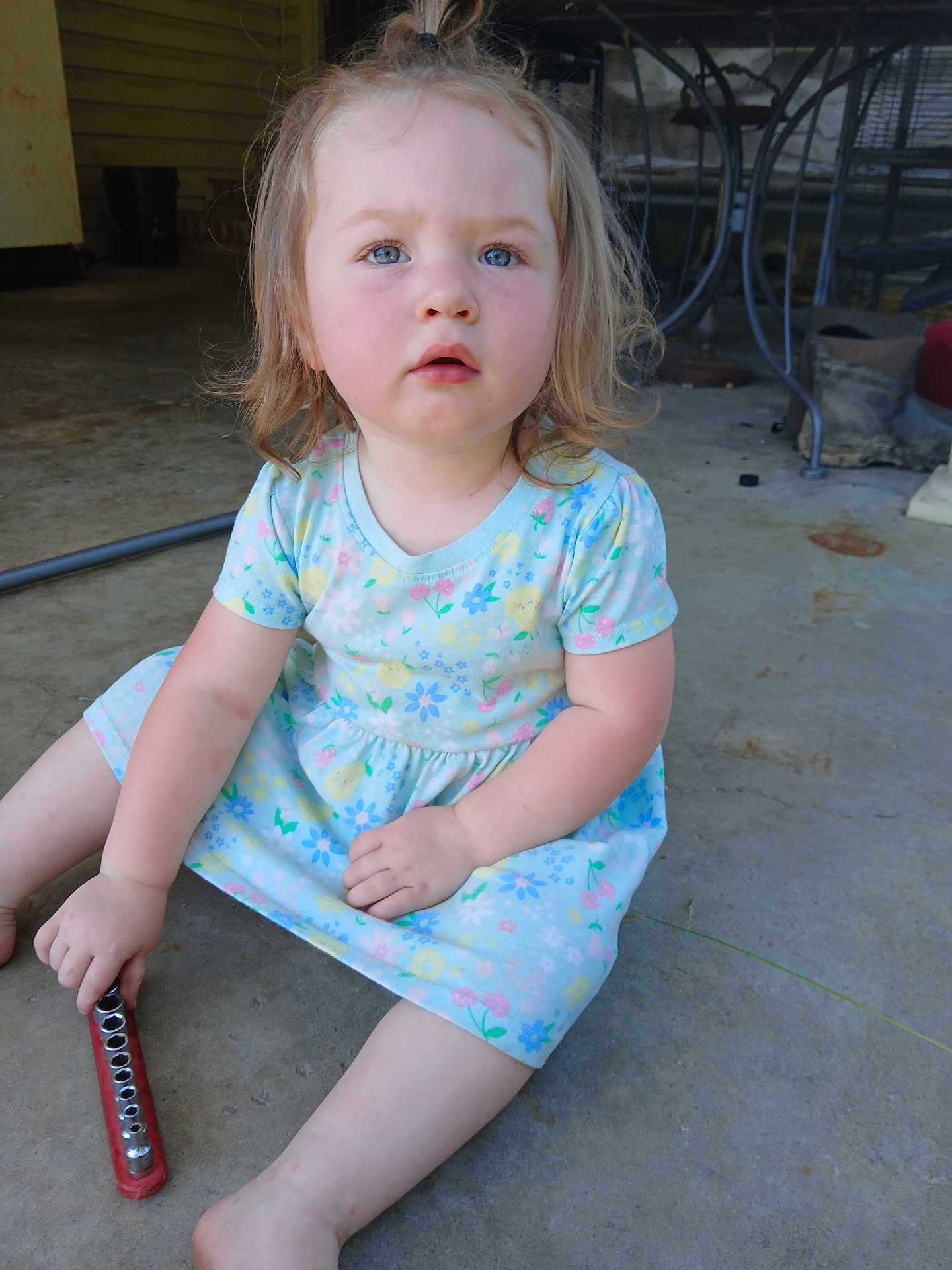 Lillian joined the competition — help win amazing prizes! toddler, child, sitting, dress, floral, concrete_floor, tool, curious, blue_eyes, hair, indoor, garage, metal, toy, expression, casual, barefoot, young_child, person, play