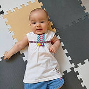 Kiona a rejoint le concours — aidez-le/la à gagner de superbes lots ! baby, baby_girl, child, childrens_clothing, colorful, cute, denim_shorts, floor, foam_mat, happy, indoor, infant, laying_down, person, play_mat, portrait, small_child, smiling, white_dress, young_child