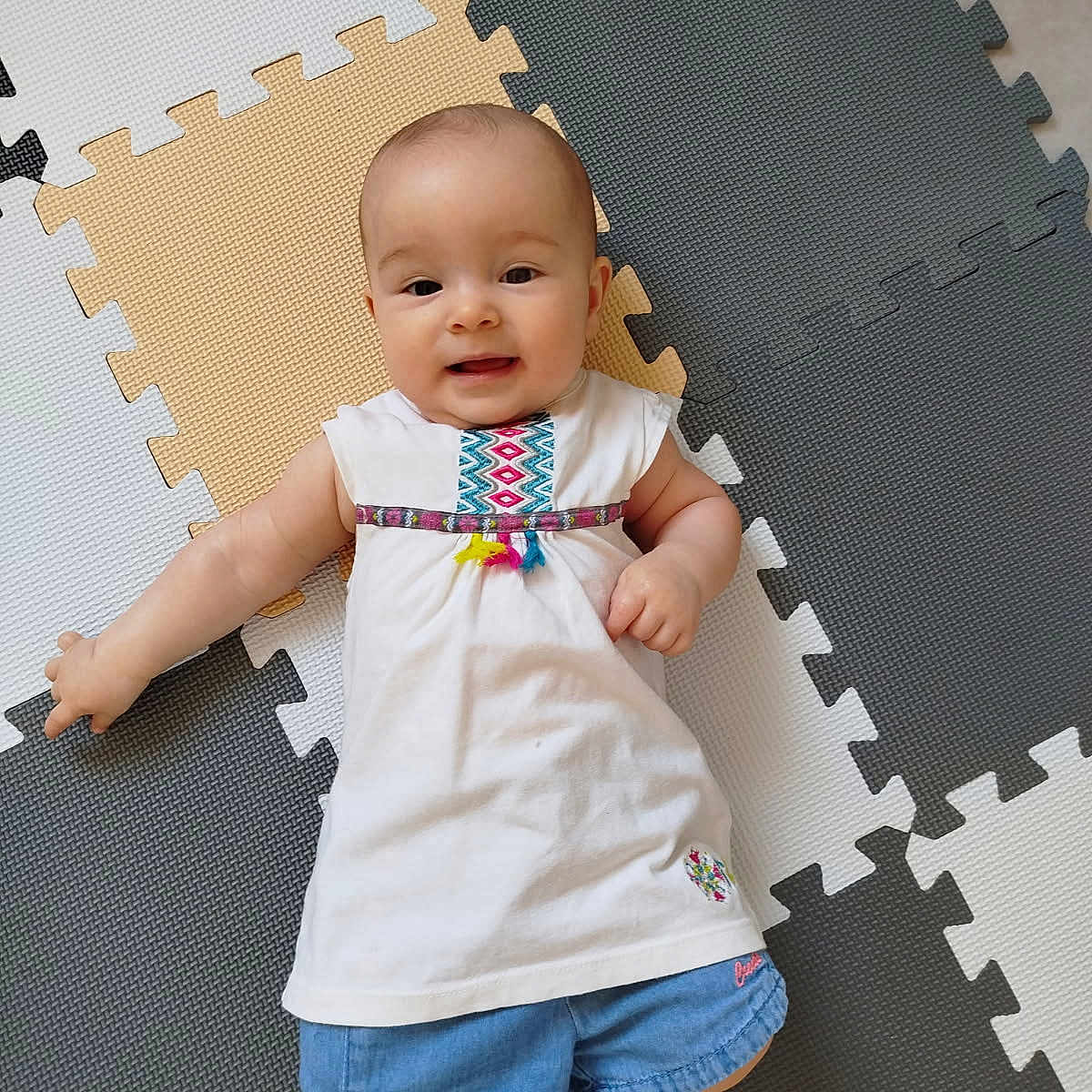 Kiona a rejoint le concours — aidez-le/la à gagner de superbes lots ! baby, baby_girl, child, childrens_clothing, colorful, cute, denim_shorts, floor, foam_mat, happy, indoor, infant, laying_down, person, play_mat, portrait, small_child, smiling, white_dress, young_child