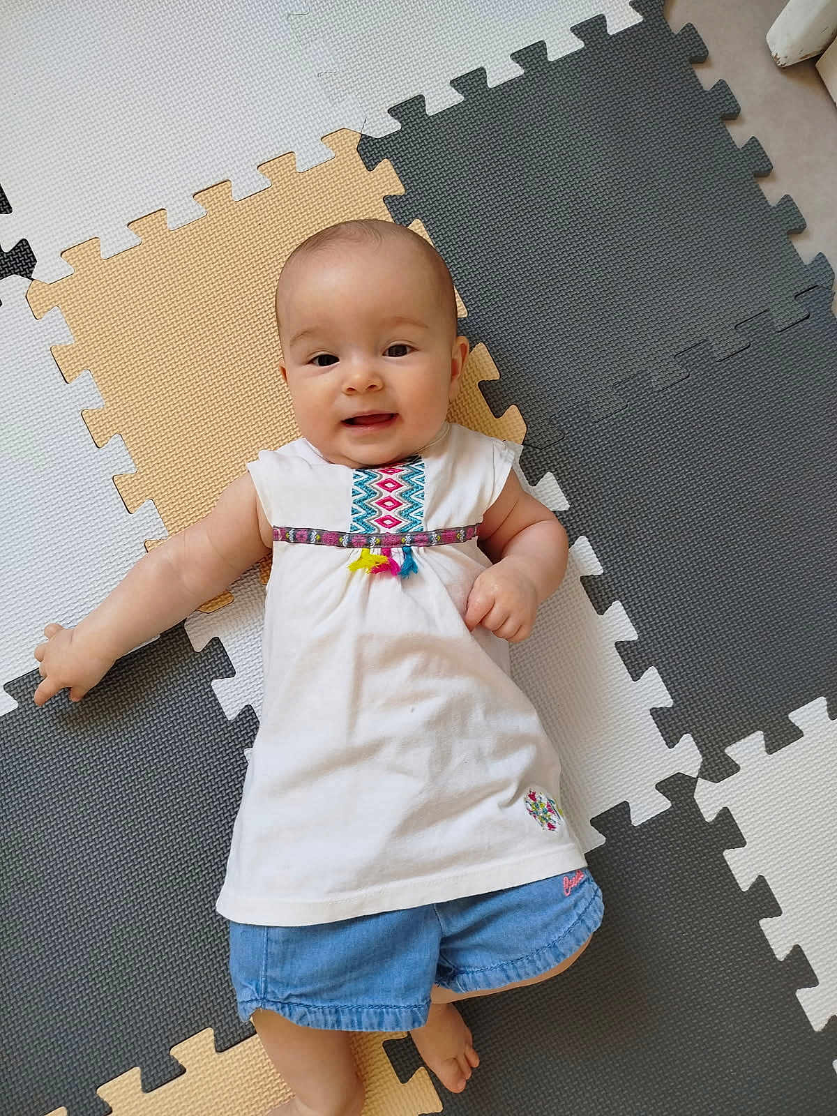 Kiona a rejoint le concours — aidez-le/la à gagner de superbes lots ! baby, baby_girl, child, childrens_clothing, colorful, cute, denim_shorts, floor, foam_mat, happy, indoor, infant, laying_down, person, play_mat, portrait, small_child, smiling, white_dress, young_child