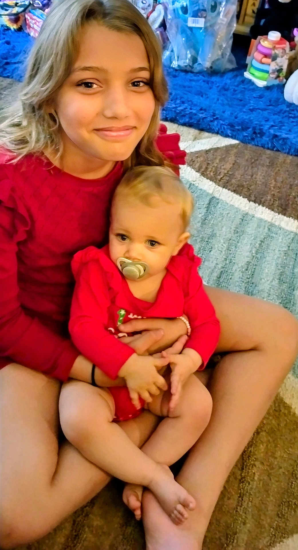 Savannah is registered to the contest to win money with this photo: child, baby, sibling, pacifier, rug, carpet, toys, indoor, sitting, smile, red_clothing, arms, legs, portrait, family, play_room, warm_lighting, cozy, holding, happy