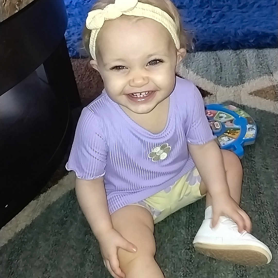 Savannah is registered to the contest to win money with this photo: bookshelf, carpet, child, coffee_table, happy, headband, indoor, living_room, playtime, portrait, purple_shirt, rug, shelf, shoe, sitting, smile, sock, toddler, toy, toy_station