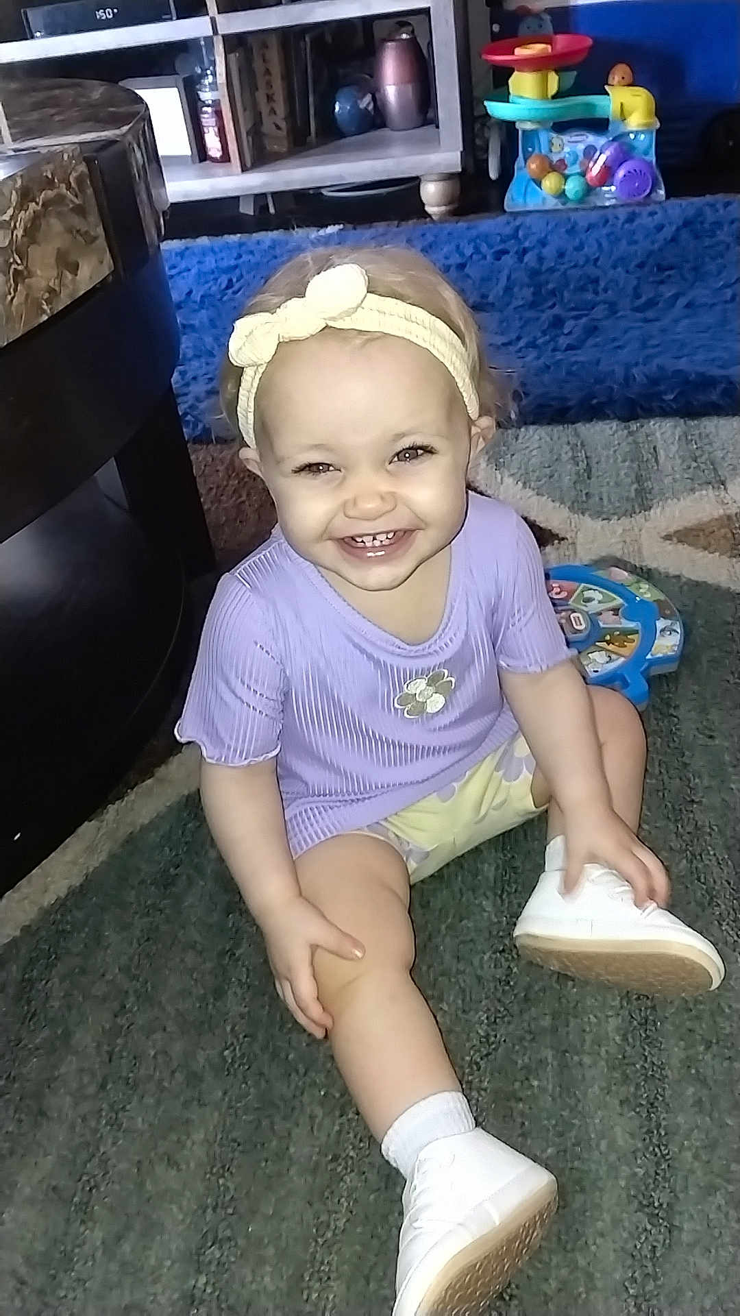 Savannah is registered to the contest to win money with this photo: child, toddler, smile, headband, purple_shirt, shoe, sock, carpet, rug, toy, toy_station, living_room, coffee_table, bookshelf, shelf, playtime, happy, sitting, portrait, indoor
