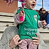 toddler, child, person, pom_pom, green_shirt, pink_pants, sneakers, knitted_sweater, bleachers, outdoor, woman, sunglasses, metal_roof, fence, excitement, smile, hands, sports_event, stadium, casual_clothing