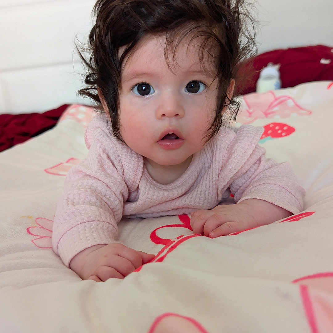 Reina joined the competition — help win amazing prizes! baby, child, infant, face, hair, bed, blanket, pink, curious, wide_eyes, lying_down, indoors, pajamas, cute, young, portrait, person, hands, expression, soft