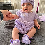 Elizabeth is registered to the contest to win money with this photo: baby, child, knit_hat, knit_dress, booties, hand_holding, indoor, gray_blanket, smiling, cute, sitting, person, soft_lighting, cozy, purple, baby_clothing, happy, infant, portrait, home
