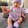 baby, child, knit_hat, knit_dress, booties, hand_holding, indoor, gray_blanket, smiling, cute, sitting, person, soft_lighting, cozy, purple, baby_clothing, happy, infant, portrait, home