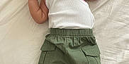 Alaska is registered to the contest to win money with this photo: baby, infant, child, white_tank_top, green_pants, cargo_pants, barefoot, lying_down, bed_sheet, curious_expression, small_hands, indoors, casual_clothing, portrait, young_child, skin, face, head, resting, cute