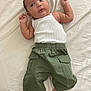 baby, infant, child, white_tank_top, green_pants, cargo_pants, barefoot, lying_down, bed_sheet, curious_expression, small_hands, indoors, casual_clothing, portrait, young_child, skin, face, head, resting, cute