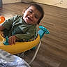 toddler, baby_walker, child, smiling, indoor, floor, wooden_floor, toy, clothing, green_shirt, face, happy, person, young_child, playful, home, room, electric_outlet, wall, cable