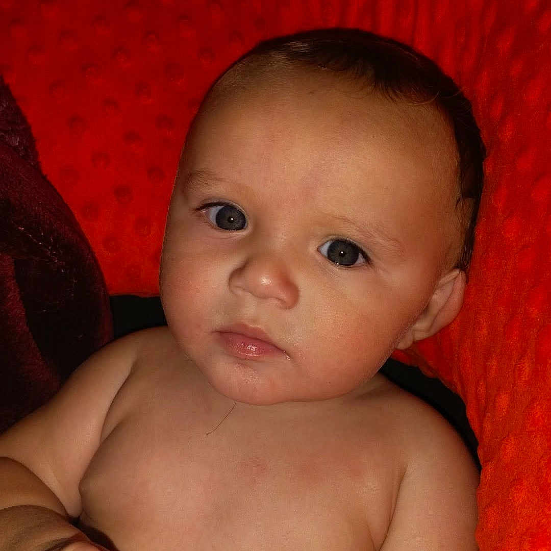 Issac is registered to the contest to win money with this photo: baby, child, bottle, face, skin, hand, red_cushion, texture, indoors, portrait, sitting, infant, closeup, young, cute, expression, looking, relaxed, holding, comfort
