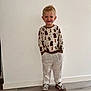 child, boy, smiling, standing, sweater, animal_print, pants, striped_pants, sneakers, hands_in_pockets, indoor, wooden_floor, white_wall, blond_hair, casual_clothing, happy, portrait, full_body, cute, young