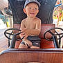 toddler, child, boy, cap, shorts, shirtless, steering_wheel, ride_on_car, amusement_park, playful, smiling, seat, outdoor, sunlight, fun, toy, vehicle, happy, summer, casual