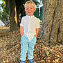 child, boy, smiling, outdoor, tree, leaves, grass, sandals, pants, shirt, blonde_hair, standing, nature, sunlight, casual_clothing, happy, portrait, summer, daytime, forest