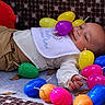 baby, child, easter_eggs, colorful, toy, playpen, smiling, lying_down, indoor, bib, pants, face, hand, soft_toys, patterned_background, play, infant, happy, plastic_eggs, cute