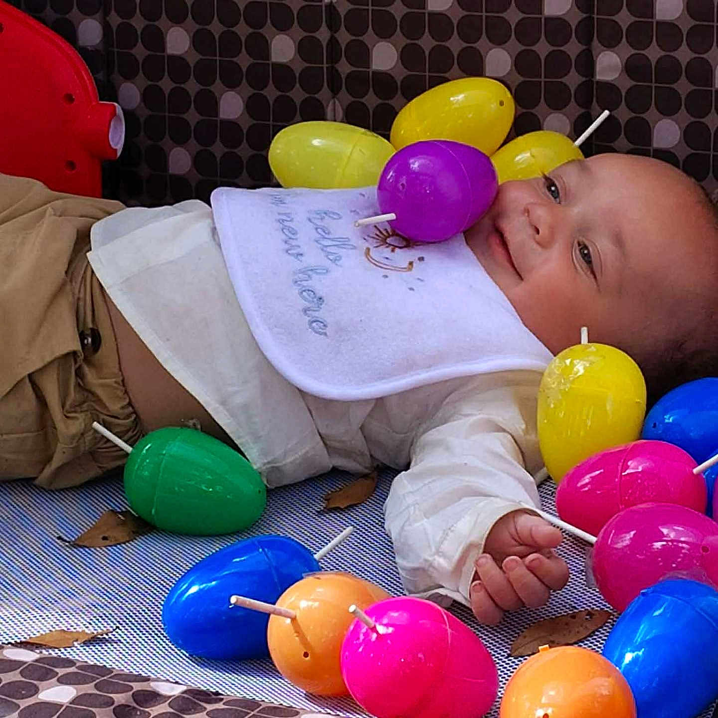 Kaior is registered to the contest to win money with this photo: baby, child, easter_eggs, colorful, toy, playpen, smiling, lying_down, indoor, bib, pants, face, hand, soft_toys, patterned_background, play, infant, happy, plastic_eggs, cute
