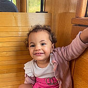 Lyah a rejoint le concours — aidez-le/la à gagner de superbes lots ! toddler, child, smiling, curly_hair, pink_skirt, cardigan, wooden_bench, indoor, happy, cute, portrait, clothing, person, face, sitting, baby, fashion, expression, young_child, casual