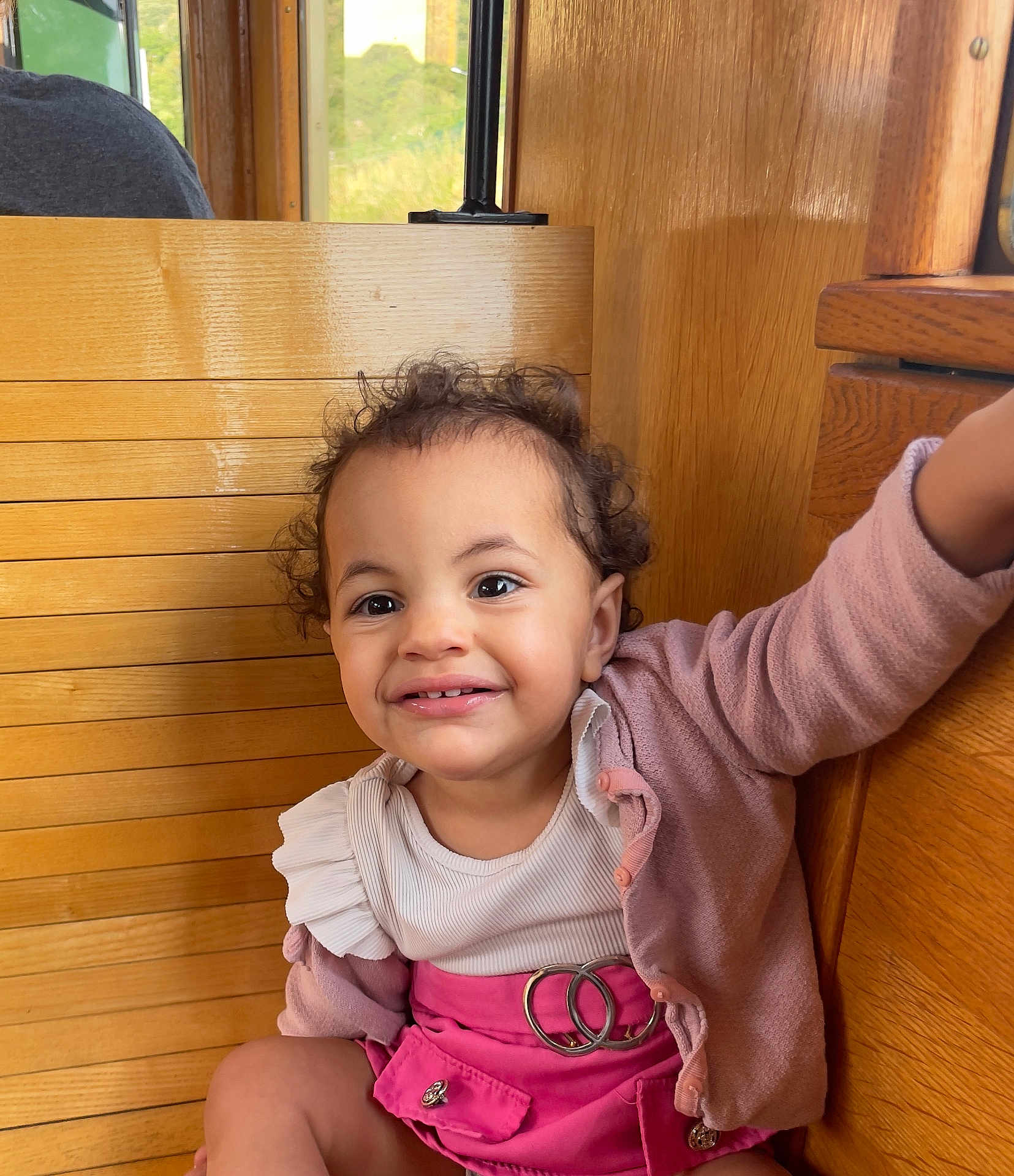Lyah a rejoint le concours — aidez-le/la à gagner de superbes lots ! toddler, child, smiling, curly_hair, pink_skirt, cardigan, wooden_bench, indoor, happy, cute, portrait, clothing, person, face, sitting, baby, fashion, expression, young_child, casual