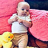 baby, infant, plush_toy, duck, pink_cushion, white_shirt, patterned_pants, child, soft_toy, cute, indoors, relaxed, seated, toddler, furniture, cozy, portrait, small_child, toy, comfort