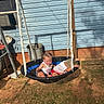 air_conditioner, blue_wall, bucket, child, clothing, daylight, dirt, fun, grass, metal, outdoor, person, playground, recreation, shadow, summer, sunlight, swing, toddler, wood