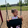 child, girl, swing, playground, outdoor, summer, park, chains, woodchips, grass, trees, building, casual_clothing, daylight, curly_hair, expression, seat, holding, young, nature