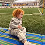 toddler, child, red_hair, gray_tracksuit, sneakers, indoor_soccer_field, artificial_turf, sports, playing, mat, striped_mat, stadium_lights, people, sports_activity, seating_area, green_turf, smiling, casual_clothing, athletic_shoes, recreation