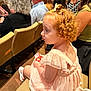 child, girl, curly_hair, red_hair, pink_dress, auditorium, seated, snack, people, adults, crowd, chair, event, indoor, side_profile, curly_hair_bow, light_skin, casual_clothing, curiosity, family