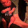 Liam is registered to the contest to win money with this photo: baby, infant, child, face, portrait, sitting, lap, adult, hand, clothing, black_outfit, onesie, text, indoor, cushion, chair, carpet, warm_lighting, red_lighting, family