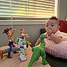 baby, toy_story, woody, jessie, buzz_lightyear, rex, toy_figures, pink_cushion, window_blinds, indoor, child, soft_texture, playtime, curious, red_bracelet, hat, lying_down, plastic_toys, colorful, home