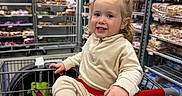 Rosalie is registered to the contest to win money with this photo: toddler, child, shopping_cart, grocery_store, bread, shelf, smile, curly_hair, clothing, indoor, aisle, person, retail, food, packaged_food, happy, casual, red, beige, store