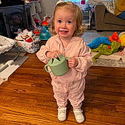 Rosalie is registered to the contest to win money with this photo: toddler, child, pink_outfit, toy, watering_can, wooden_floor, living_room, couch, person, smile, curly_hair, indoor, playtime, shoes, blanket, stuffed_toy, carpet, window, furniture, happy