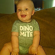 Bobby Ray is registered to the contest to win money with this photo: baby, child, smiling, onesie, dinosaur, couch, indoor, living_room, happy, sitting, cute, infant, person, face, cheerful, skin, furniture, home, young, expression