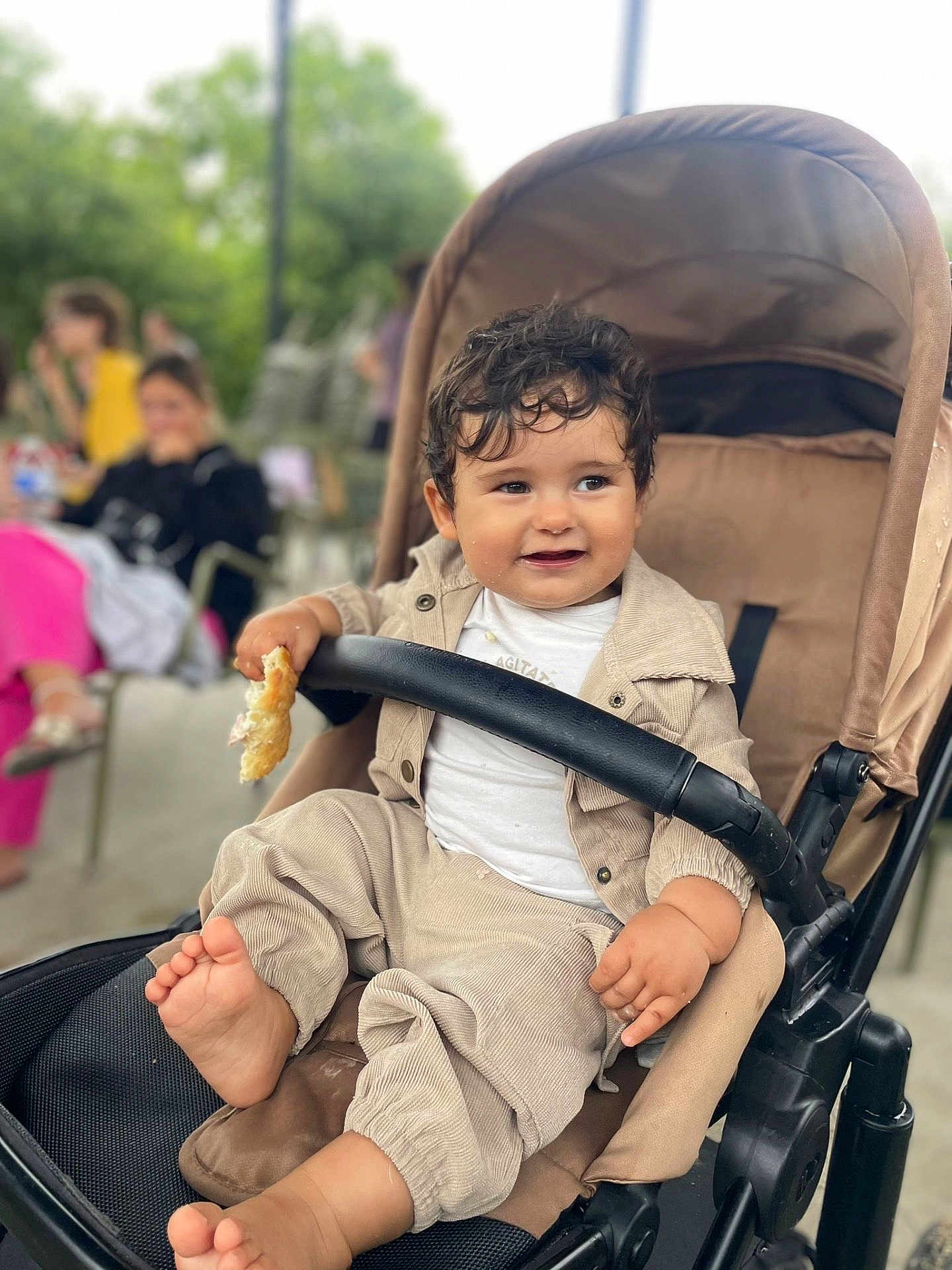 Maé a rejoint le concours — aidez-le/la à gagner de superbes lots ! toddler, child, stroller, bread, smile, curly_hair, clothing, beige_outfit, barefoot, hand, face, happy, seated, baby, outdoor, people, blurred_background, daylight, park, casual