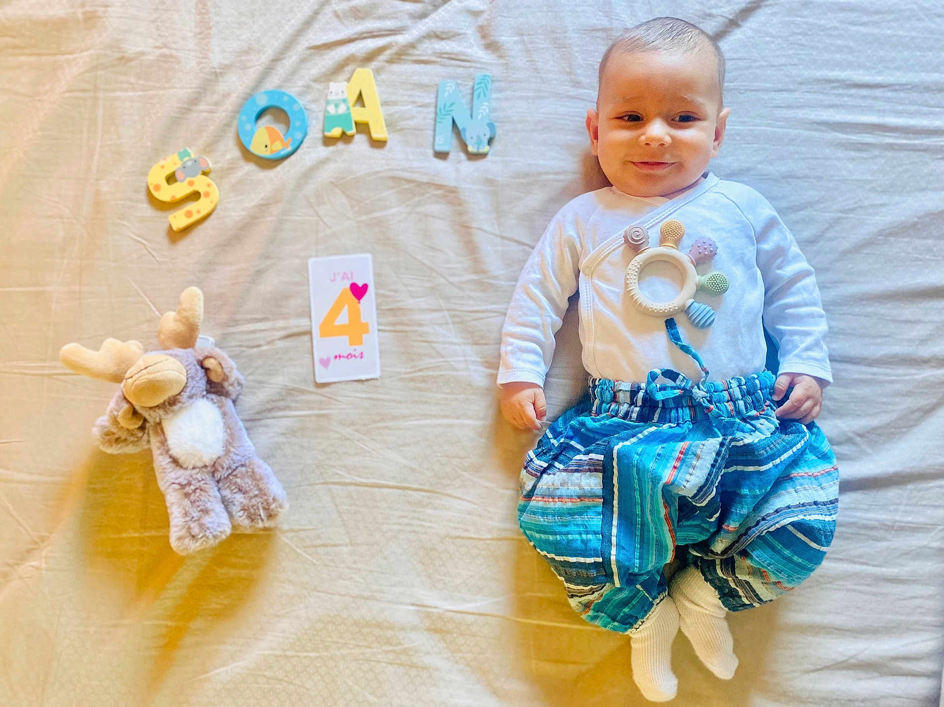 Soän participe au concours pour gagner de l'argent avec cette photo : baby, infant, child, plush_toy, moose_toy, letters, soan, card, age_card, clothing, pants, white_shirt, sock, bed, blanket, smile, face, milestone, lying_down, cute