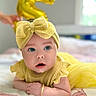 baby, child, yellow_clothing, headband, bow, bracelet, balloon, number_5, blanket, indoor, portrait, cute, infant, celebration, smiling, face, eyes, skin, hand, lying_down