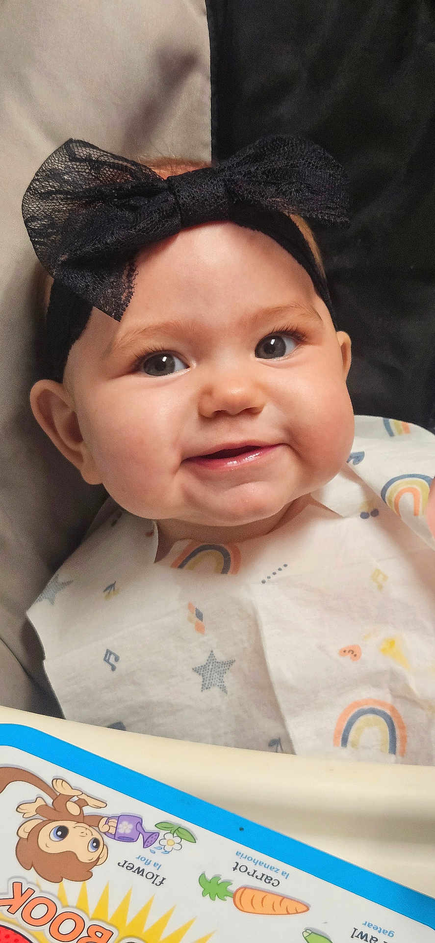 Riley is registered to the contest to win money with this photo: baby, child, smiling, headband, black_bow, bib, rainbow, stars, high_chair, book, toy, face, cheeks, cute, indoor, infant, person, happy, clothing, portrait