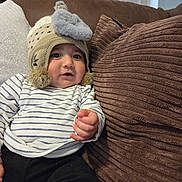 Enzo joined the competition — help win amazing prizes! baby, child, hat, knitted_hat, elephant, plush, striped_shirt, couch, pillow, brown_pillow, white_pillow, indoor, cozy, soft, curious, person, infant, casual_clothing, hand, smiling