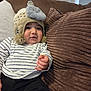 baby, child, hat, knitted_hat, elephant, plush, striped_shirt, couch, pillow, brown_pillow, white_pillow, indoor, cozy, soft, curious, person, infant, casual_clothing, hand, smiling
