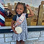 child, birthday, girl, smile, curly_hair, dress, sash, tiara, bracelet, purse, stone_wall, window, reflection, outdoor, celebration, happy, person, fashion, casual, daytime
