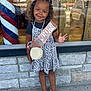 child, birthday, sash, smile, curly_hair, dress, leopard_print, sandals, sidewalk, stone_wall, window, reflection, car, outdoor, sunny, happy, person, accessory, bag, celebration