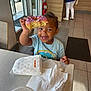 child, toddler, donut, food, table, indoor, smile, happy, person, clothing, napkin, drink, cup, straw, restaurant, counter, floor, window, chair, bag