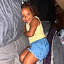 child, smiling, bed, pillow, curly_hair, yellow_tank_top, blue_shorts, happy, indoor, cozy, person, cute, toddler, skin, face, furniture, relaxed, playful, home, resting