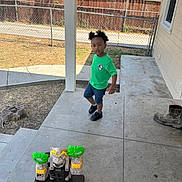 Jonnathan joined the competition — help win amazing prizes! toddler, child, green_shirt, denim_shorts, black_shoes, toy_castle, concrete_porch, boot, fence, yard, grass, tree, house, window, sunlight, outdoor, sidewalk, curious, standing, daytime